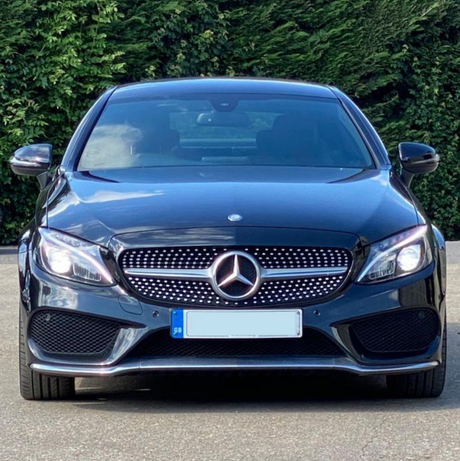 A black Mercedes-Benz car parked on a paved surface, facing forward, surrounded by green foliage. The front grille features the iconic emblem, with the license plate below.