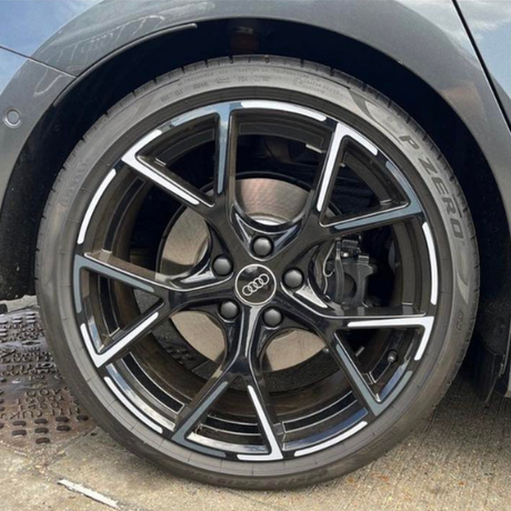 A black Audi car wheel with intricate silver spokes is parked on a concrete surface. The tire reads "P ZERO." The Audi logo is visible on the wheel center.