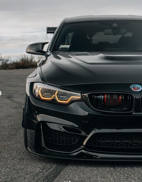 A sleek black car with glowing headlights is parked on a paved surface, with a cloudy sky in the background. The car's design is sporty and aggressive.