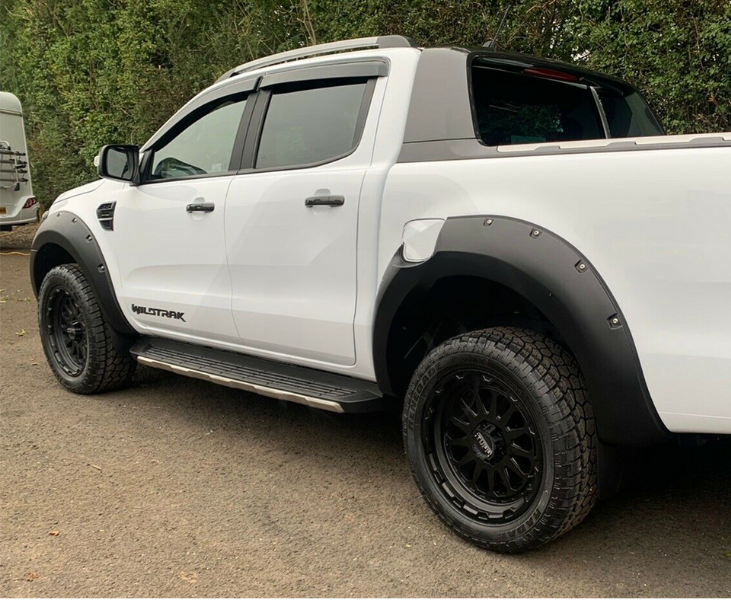 White pickup truck, parked, features black wheel arches and step bars; "WILDTRAK" text on its side. It's situated on an asphalt surface, bordered by dense green foliage.