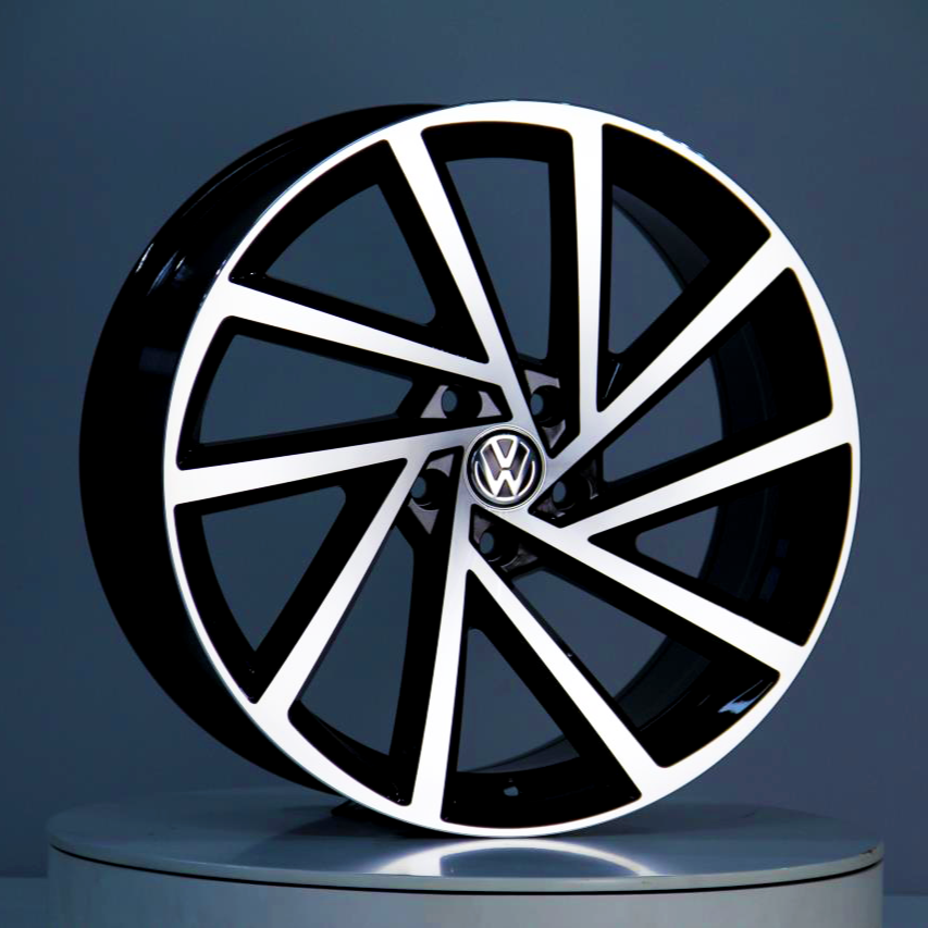 A black and silver car wheel with a geometric spoke design, displaying a Volkswagen logo in the center, is positioned upright on a reflective surface within a gray environment.