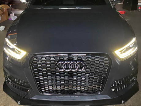 A black car with illuminated headlights, featuring a distinctive honeycomb grille with a logo. The vehicle is parked indoors, surrounded by various items and boxes.