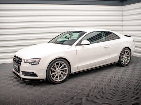 A white Audi coupe is parked, showcasing its sleek design with silver alloy wheels, in a well-lit room with textured gray flooring and horizontal white paneling on the walls.