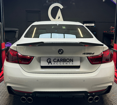 A white BMW 435d with a custom spoiler is parked on a vehicle lift inside an automotive workshop. The license plate reads "CARBON ACCENTS" with a logo above.