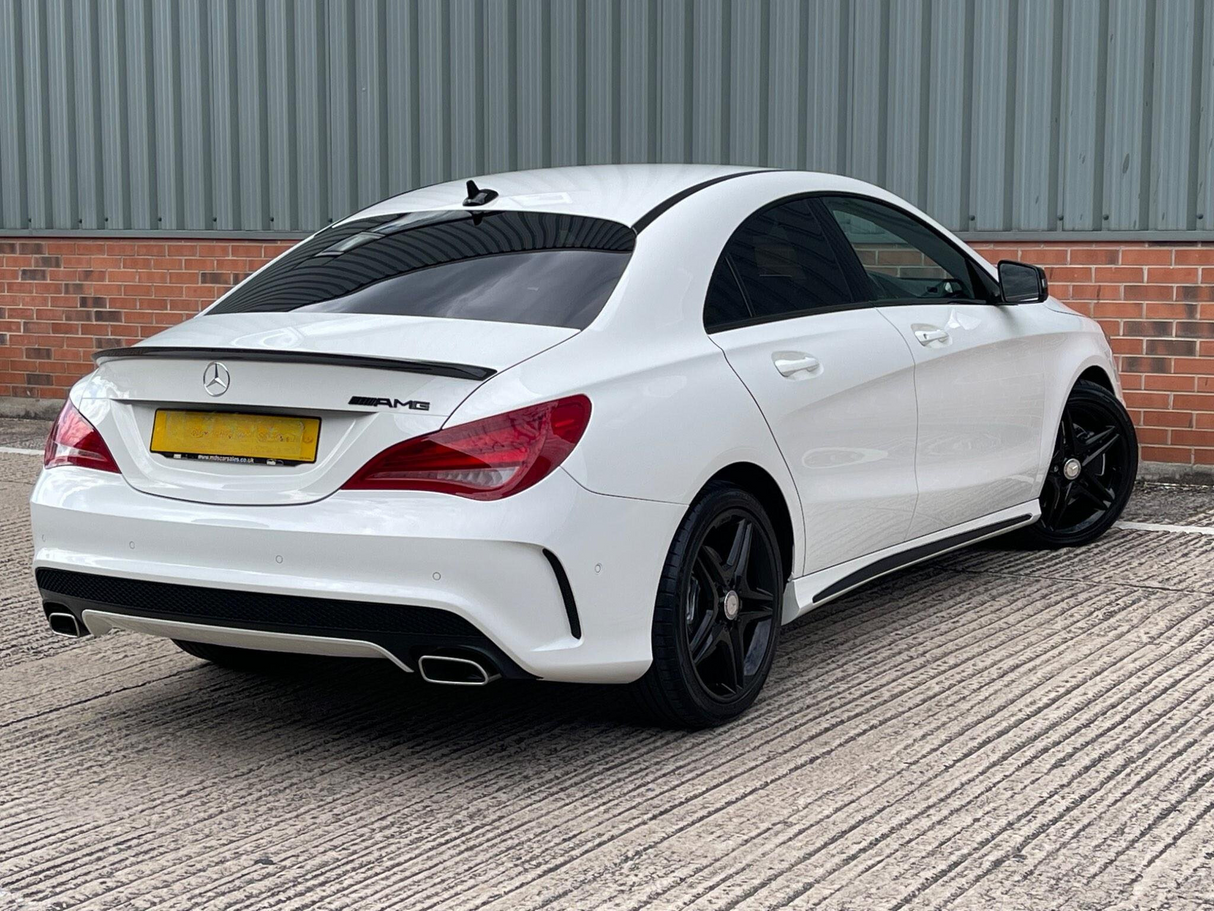 A white Mercedes-Benz AMG car is parked on a textured concrete surface beside a brick and corrugated metal wall. The car's license plate text is unclear. Black wheels and a rear spoiler enhance its sporty look.