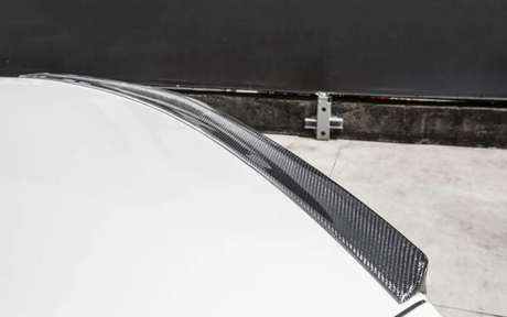 A carbon fiber car spoiler on a white vehicle is shown close-up, situated in an outdoor setting near a concrete wall and metal barrier post.