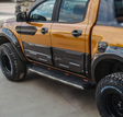 A modified pickup truck, parked, displaying a shiny orange and black exterior with large off-road tires labeled "BFGoodrich" and distinct side steps; situated on a paved surface.