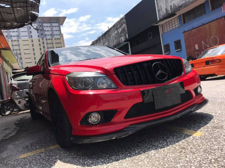 Red car parked on a street with a reflective black grille and black wheels. Surrounded by urban buildings and motorcycles under a partly cloudy sky. The car is positioned diagonally.