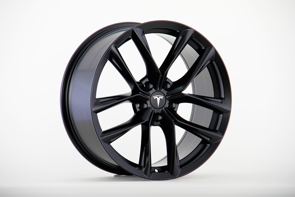 A sleek black alloy wheel is displayed upright, featuring a multi-spoke design with the Tesla logo at its center, set against a plain white background.