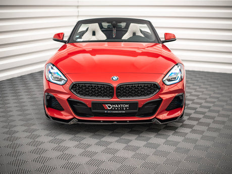 A red convertible sports car sits stationary on a patterned floor within a white-walled room. Its front grille prominently displays the BMW logo. Text on the bumper reads "MAXTON design."