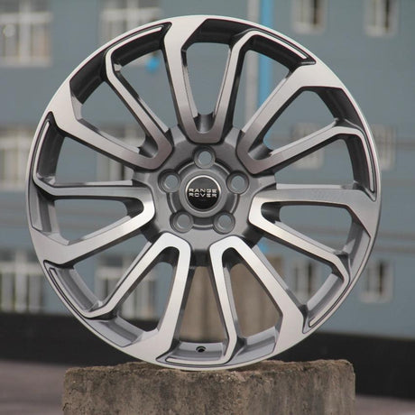 A silver alloy car wheel with intricate spoke design stands upright on a concrete block. The central hub features the text "RANGE ROVER." In the background, blurred urban buildings are visible.