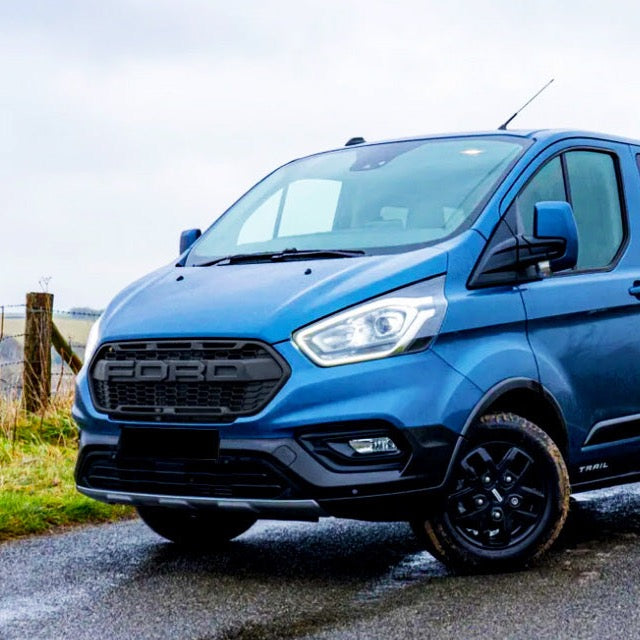 A blue van is parked on a wet rural road. The grille displays "FORD," and the word "TRAIL" is visible near the rear wheel. A wooden fence is in the background.
