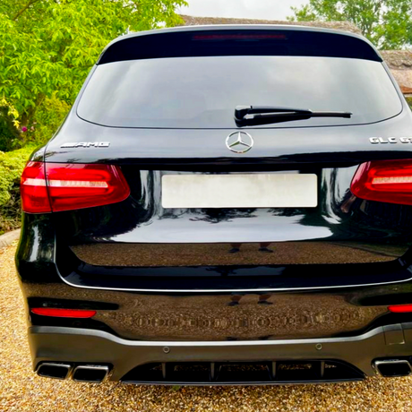 A black Mercedes-Benz GLC 63 AMG parked on a gravel driveway, surrounded by green trees. The rear of the car features four exhaust pipes and red taillights.