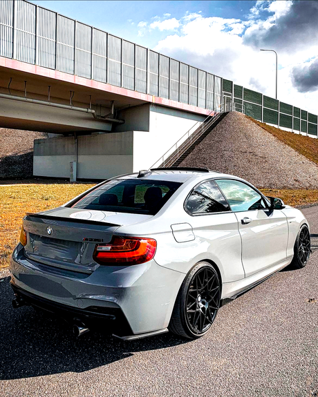 A gray BMW sports car is parked on a road beneath a large bridge with metal railings, near grassy embankments under a partly cloudy sky.
