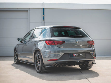 A grey SEAT Leon car is parked on a concrete surface near a white building. The rear features a Maxton Design diffuser and visible dual exhausts.