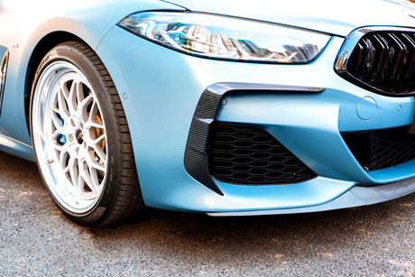 A close-up of a blue car's front, featuring a sleek headlight, sporty grille, and detailed alloy wheel. The vehicle is parked on a paved surface.