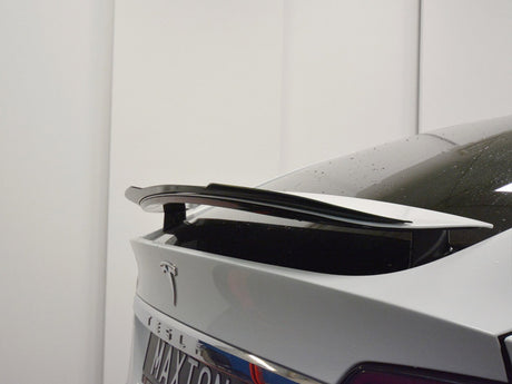 A car's rear end features a black spoiler on a white polished surface, with "MA-XTON" visible on the license plate. The background is a plain white wall.
