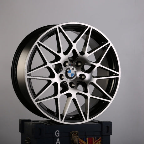 A BMW car rim with an intricate, geometric design stands upright on a dark blue box emblazoned with the letters "GA" and a partial Union Jack in a plain gray environment.