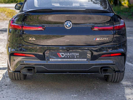 A black BMW X4 M40i is parked on a gravel surface. The license plate reads "MAXTON design." The car's rear includes dual exhausts and prominent taillights.