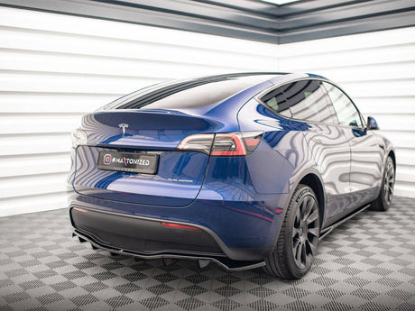 A blue Tesla Model Y, displaying a "DUAL MOTOR" badge, is parked in a well-lit, modern garage with checkered flooring. The license plate reads: "@#MAATONIZED YELRAX.COM".