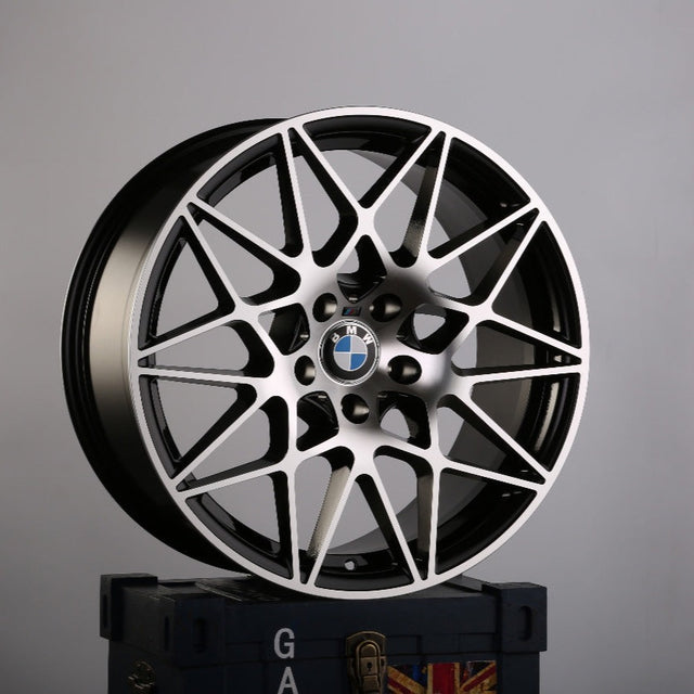 A BMW wheel with intricate multi-spoke design rests atop a dark-blue crate, featuring a faint Union Jack pattern and partial text "GA" visible, against a plain grey background.
