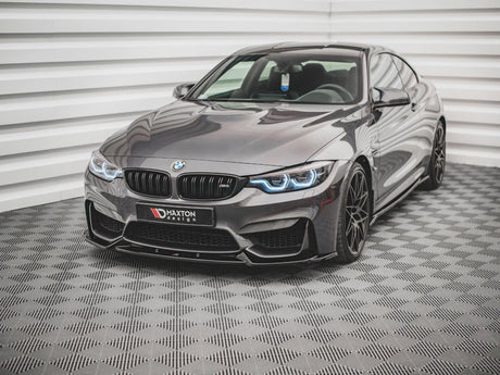 A sleek, gray BMW sports car with illuminated headlights sits on a checkered floor in a bright, white-paneled room. A license plate reads: "MAXTON design."