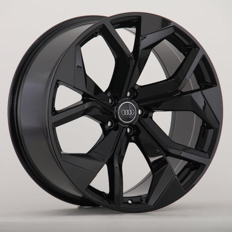 A black, glossy alloy wheel with a dynamic, angular spoke design. The Audi logo is at its center. The wheel is set against a plain, light gray background.