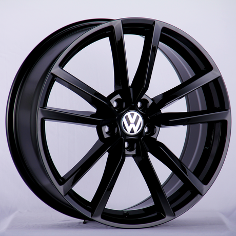 A black car wheel rim with a glossy finish stands upright. The center features a logo with a "VW" emblem. The background is a plain light surface.