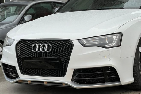 The white car features a prominent hexagonal grille with four interlinked rings, parked next to another vehicle in an indoor setting. The headlight design is sleek and modern.