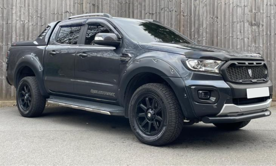 A dark gray pickup truck is parked on a paved surface. It features large black wheels and a prominent front grille. In the background, there is a wooden fence.