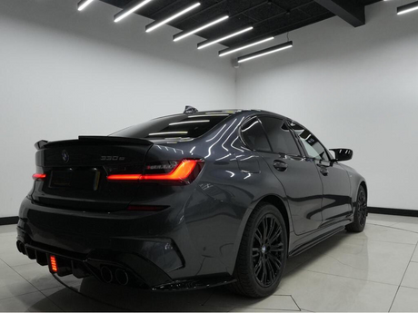 A sleek, dark-colored BMW 330e parked indoors with bright, linear ceiling lights reflecting on its polished surface. The rear of the car features illuminated taillights and visible exhaust pipes.