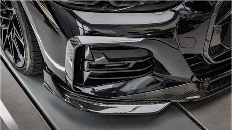 A black car with a carbon fiber front bumper detail is parked in a garage, reflecting nearby objects and lights on its glossy surface.