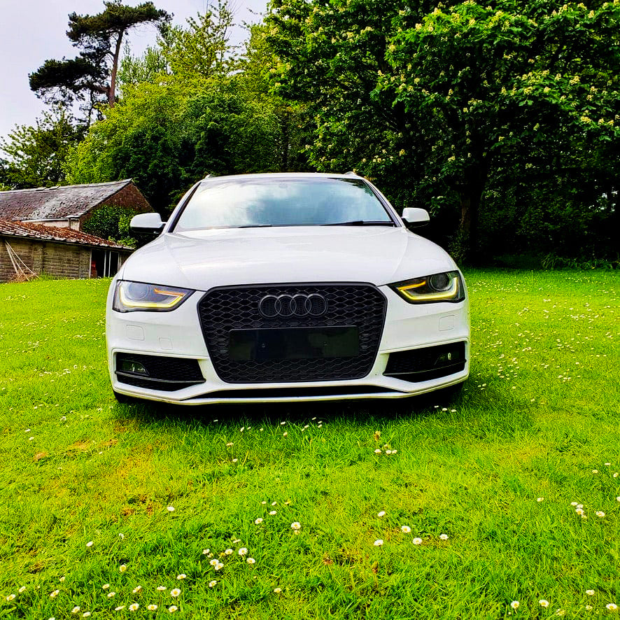 A white Audi car is parked on a grassy field, surrounded by lush green trees and a rustic building in the background.