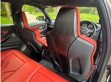 Car interior with red leather seats and carbon fiber backrests, showcasing a sleek dashboard and steering wheel, viewed from the backseat. Greenery is visible through the window.