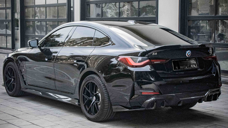 A black BMW i4 parked on a cobblestone driveway outside a modern building with large windows. The car's design features sleek lines and tinted windows, with badges displaying "BMW," "i4," and "M50".