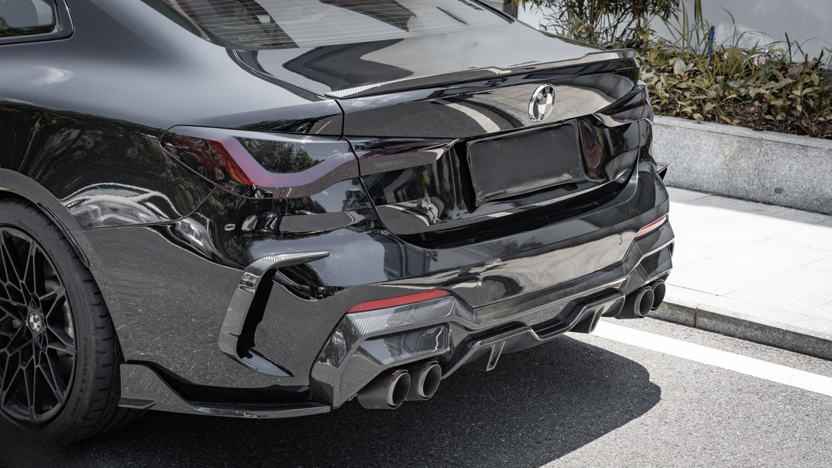 A black sports car is parked on a street, featuring a sleek rear design, quad exhaust pipes, and a BMW logo. Surrounding foliage lines a nearby concrete boundary.