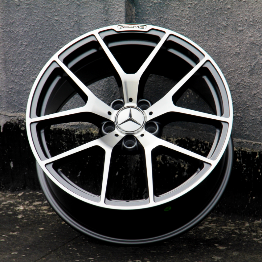 A metallic alloy wheel with a star emblem and "AMG" logo is standing upright against a textured gray wall, positioned on a dark concrete surface.