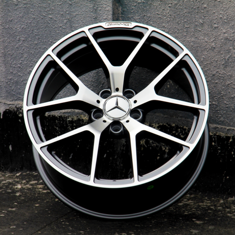 A metallic alloy wheel with a star emblem and "AMG" logo is standing upright against a textured gray wall, positioned on a dark concrete surface.