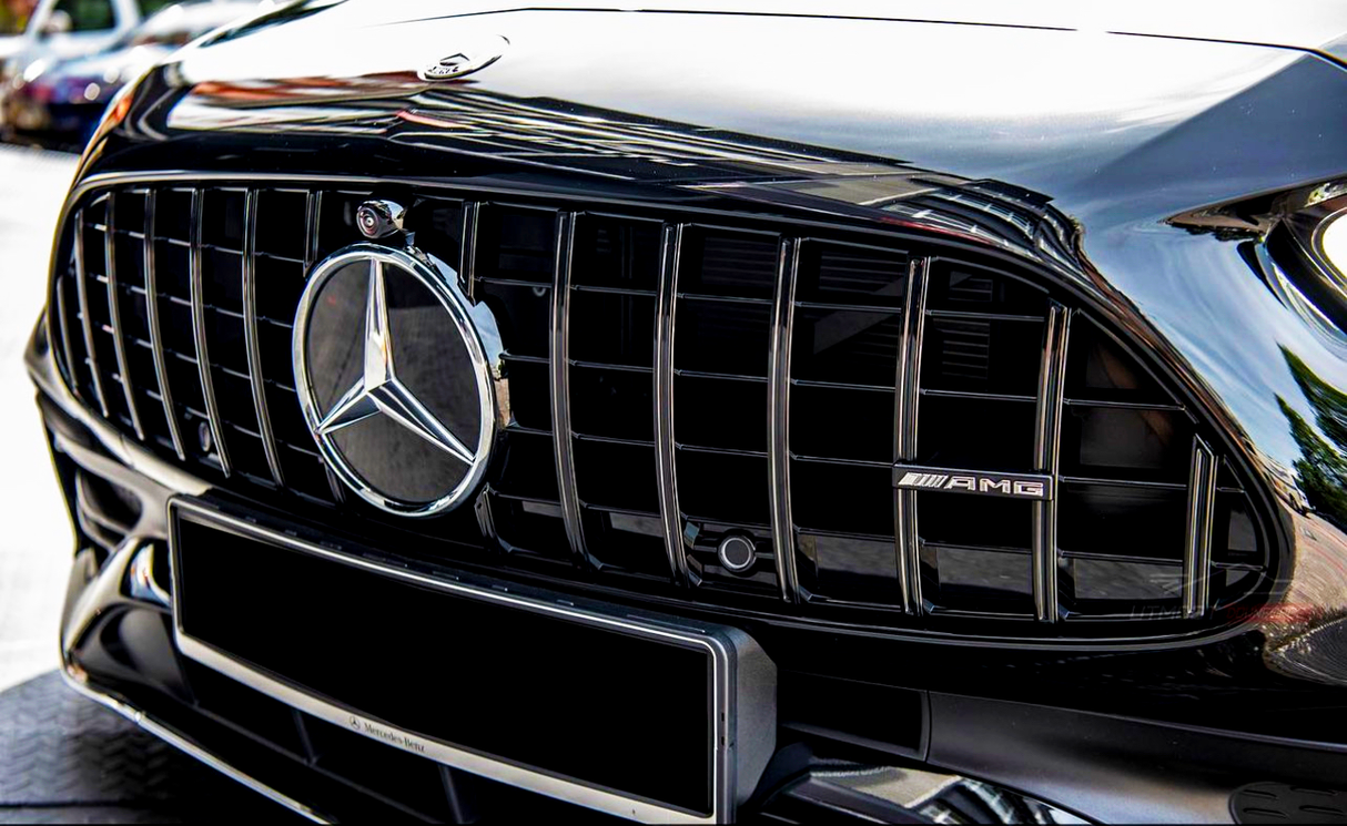 A black car grille features a prominent Mercedes-Benz logo and AMG badge. The grille has vertical slats, reflecting sunlight. Parked vehicles and a building are faintly visible in the background.