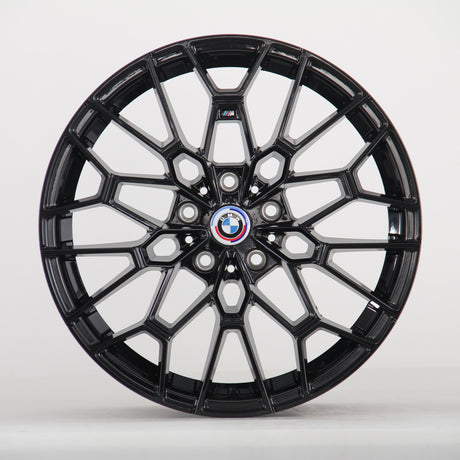 The image shows a black alloy wheel with a complex geometric design, featuring a blue, white, and red BMW logo at the center. The wheel is set against a plain white background.