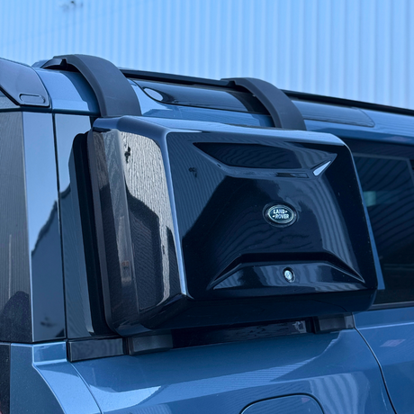 A black, angular box with the "Land Rover" logo is mounted on the rear of a blue vehicle, surrounded by a metallic rooftop in an outdoor setting.