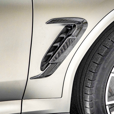 A carbon fiber air vent is attached to the side of a light-colored vehicle, located near the edge of a black tire with visible tread patterns.