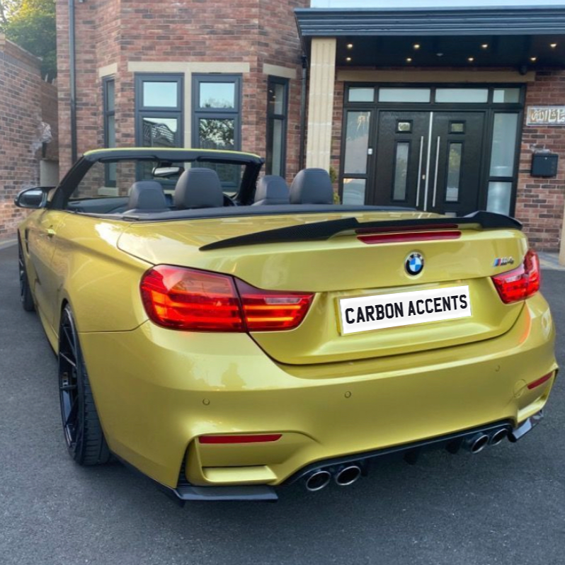 A yellow convertible car is parked in front of a brick building. The license plate reads "CARBON ACCENTS," and the car features a quad exhaust system.