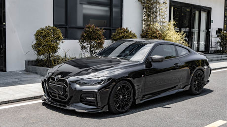 A sleek black sports car is parked on a street, illuminated by sunlight. It's positioned in front of a modern building with large windows, surrounded by decorative shrubs.