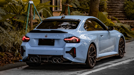 A blue sports car with distinctive tail lights and dual exhausts is parked on a road, surrounded by lush green vegetation and a staircase in the background.