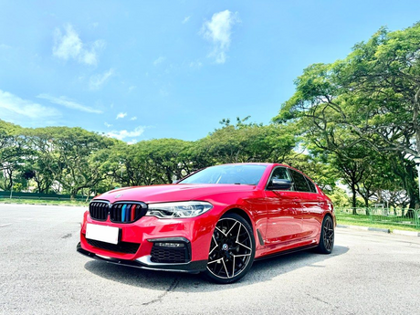 A red car is parked on a paved surface under a clear sky, surrounded by lush green trees in the background.