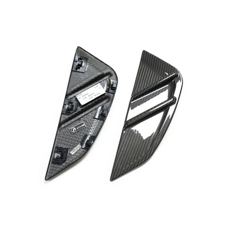 Two carbon fiber car trim pieces lie on a plain surface; one shows a glossy, ribbed exterior, while the other reveals a textured interior with mounting clips and labels.