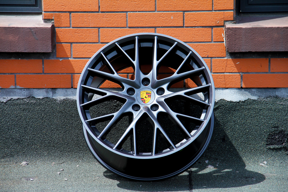 A black alloy wheel with a yellow Porsche emblem, leaning against an orange brick wall, casting a shadow on a textured ground surface.