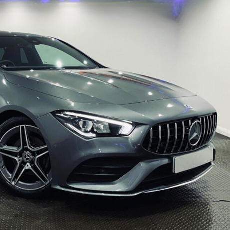 A sleek, silver luxury car is parked on a shiny floor in a brightly lit showroom, featuring a prominent front grille and stylish alloy wheels.