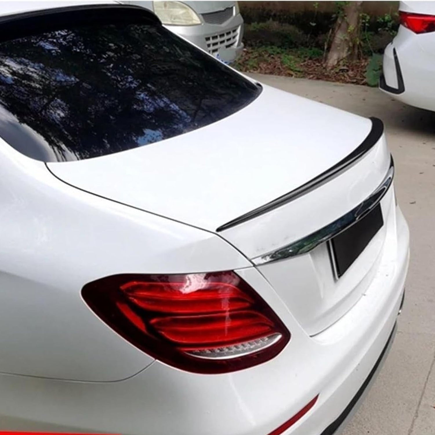 A white car is parked with visible red taillights. It has a black spoiler on the trunk. Nearby, other vehicles and trees are visible.